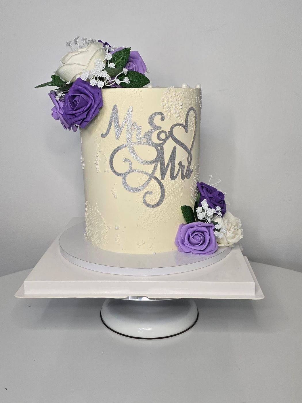 A cream wedding cake with a silver Mr and Mrs topper and purple roses by Cakes by Finest